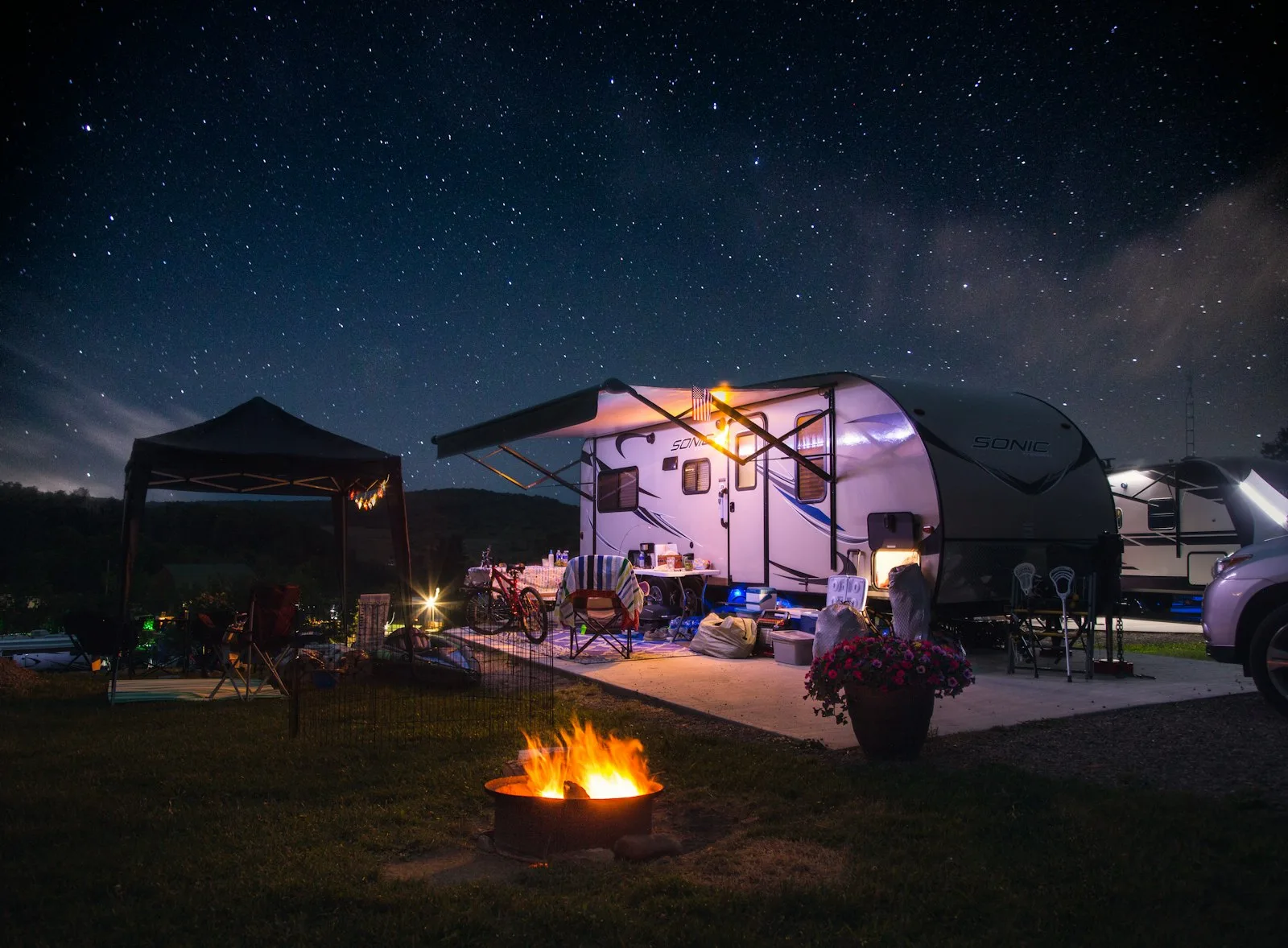 A travel trailer parked at a campsite at night with a bonfire in the foreground.