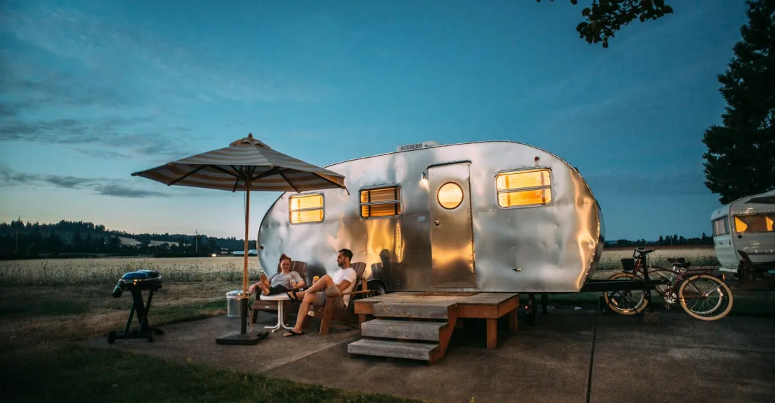 A couple sitting in chairs outside a shiny silver travel trailer at dusk.